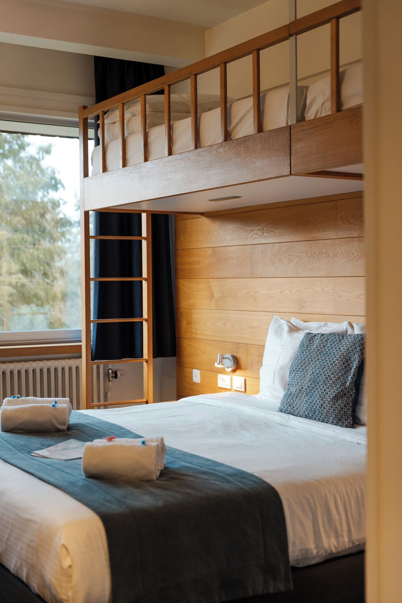Une chambre avec un lit superposé doté d’une tête de lit en bois, une table de chevet avec une lampe et une fenêtre donnant sur des arbres.