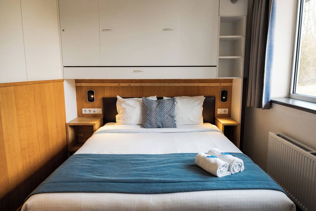 Une chambre d’hôtel avec un lit, une tête de lit en bois, du linge de lit blanc, une couverture bleue et une fenêtre avec des rideaux.