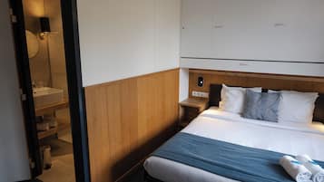 Habitación de hotel con cama, cabecera de madera, mesita de noche, lámpara de pared y baño visible a través de una puerta abierta.