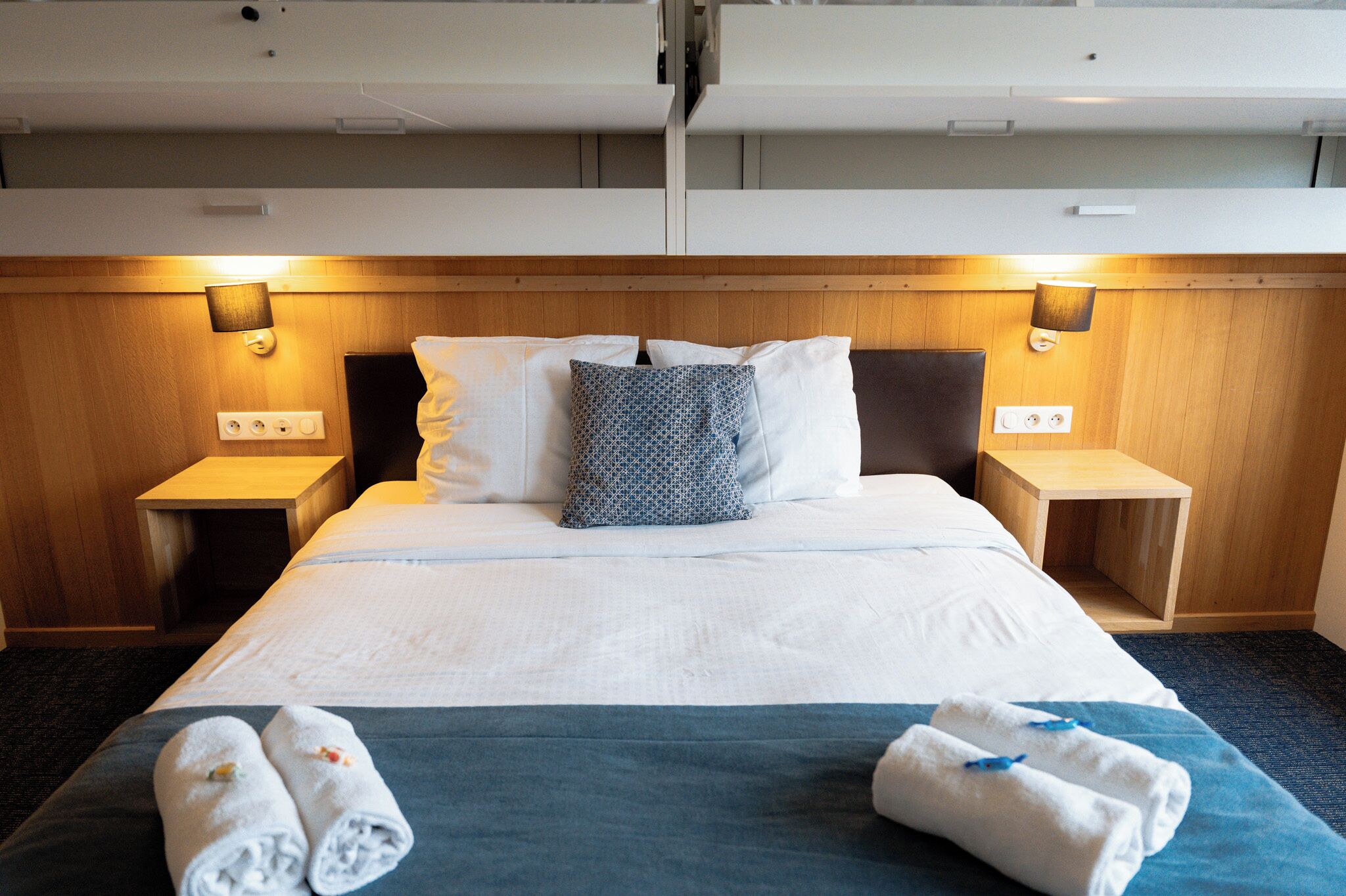 Quarto de hotel com cama, duas mesas de cabeceira, painel de cabeceira e duas luminárias fixadas na parede.