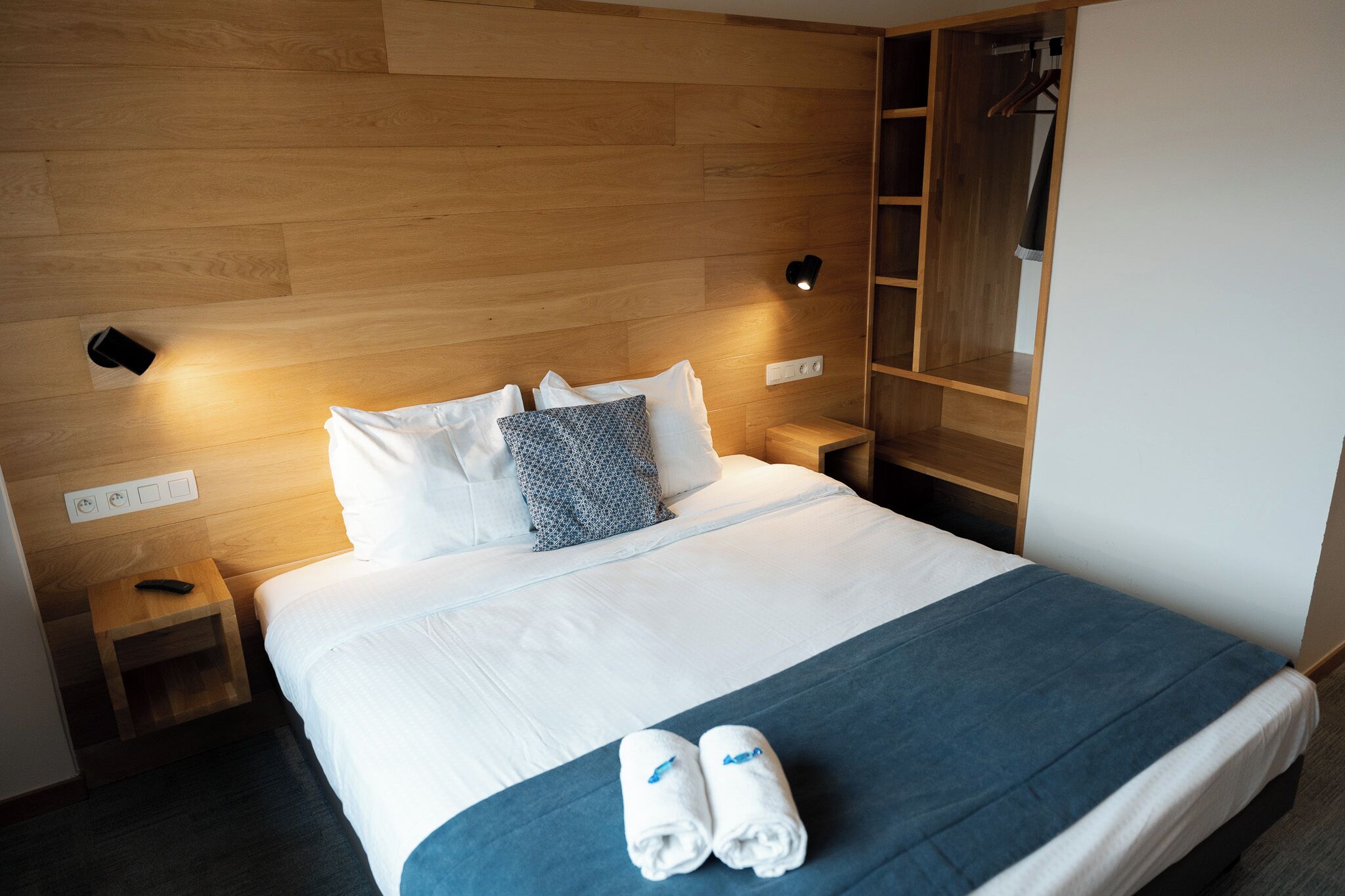 A hotel room with a wooden headboard, a bed with white linens, two bedside tables, a nightstand, a wardrobe, and a wall-mounted light fixture.