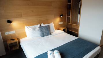A hotel room with a wooden headboard, a bed with white linens, two bedside tables, a nightstand, a wardrobe, and a wall-mounted light fixture.