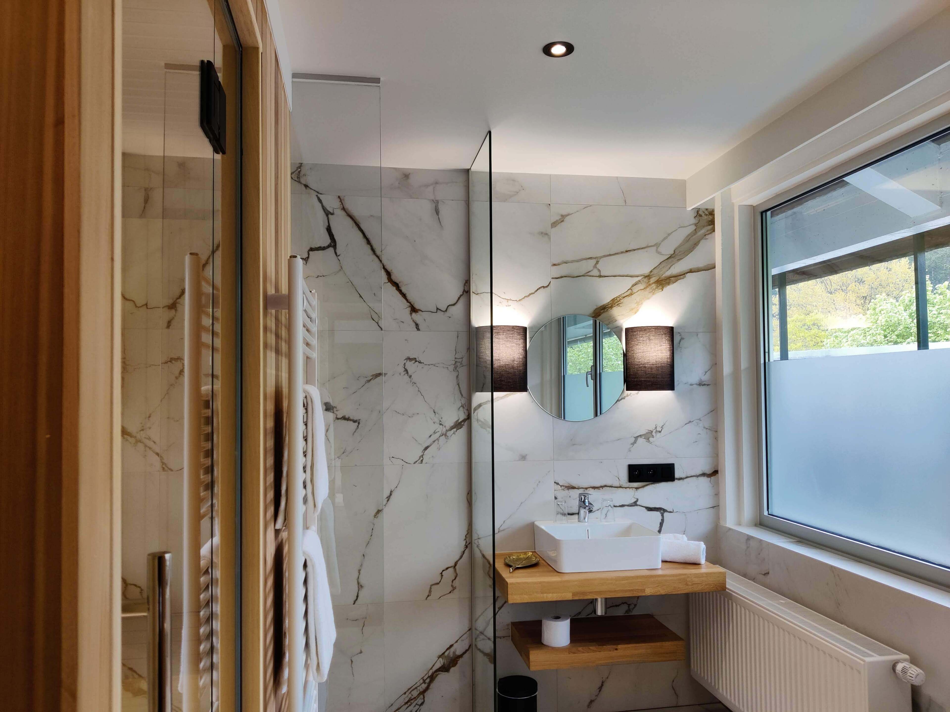 A bathroom with a marble wall, a round mirror, a white sink, a wooden shelf, and a window.