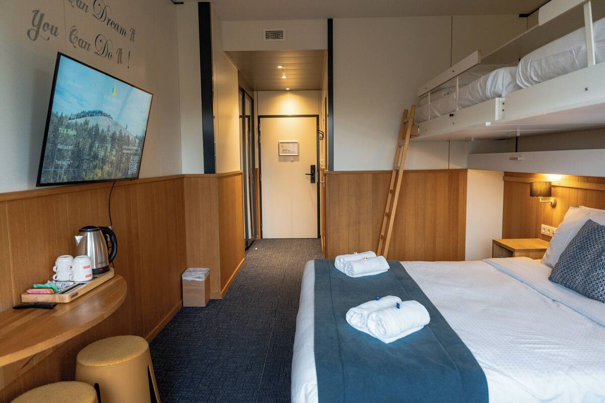 Une chambre d’hôtel avec un lit superposé, un téléviseur fixé au mur, un bureau en bois avec une bouilloire et un petit tabouret.