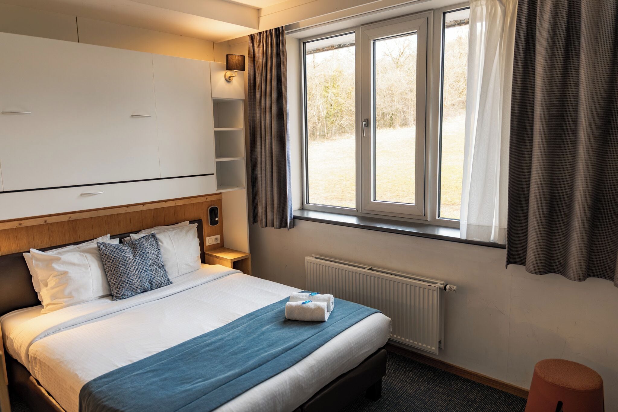 A hotel room with a bed, a window with curtains, a radiator, and a wardrobe.