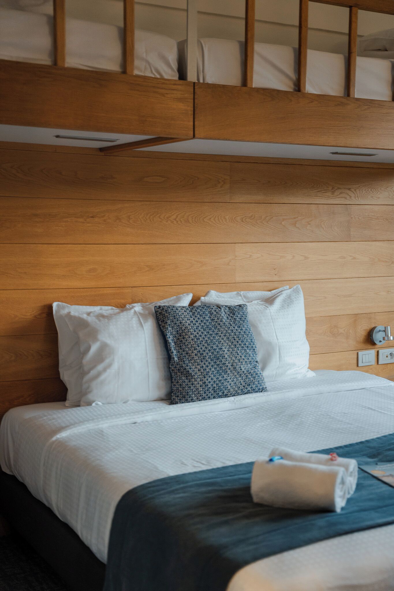 A hotel room with a wooden headboard, a bed with white and patterned pillows, a dark blue bedspread, and a rolled towel on the bed.