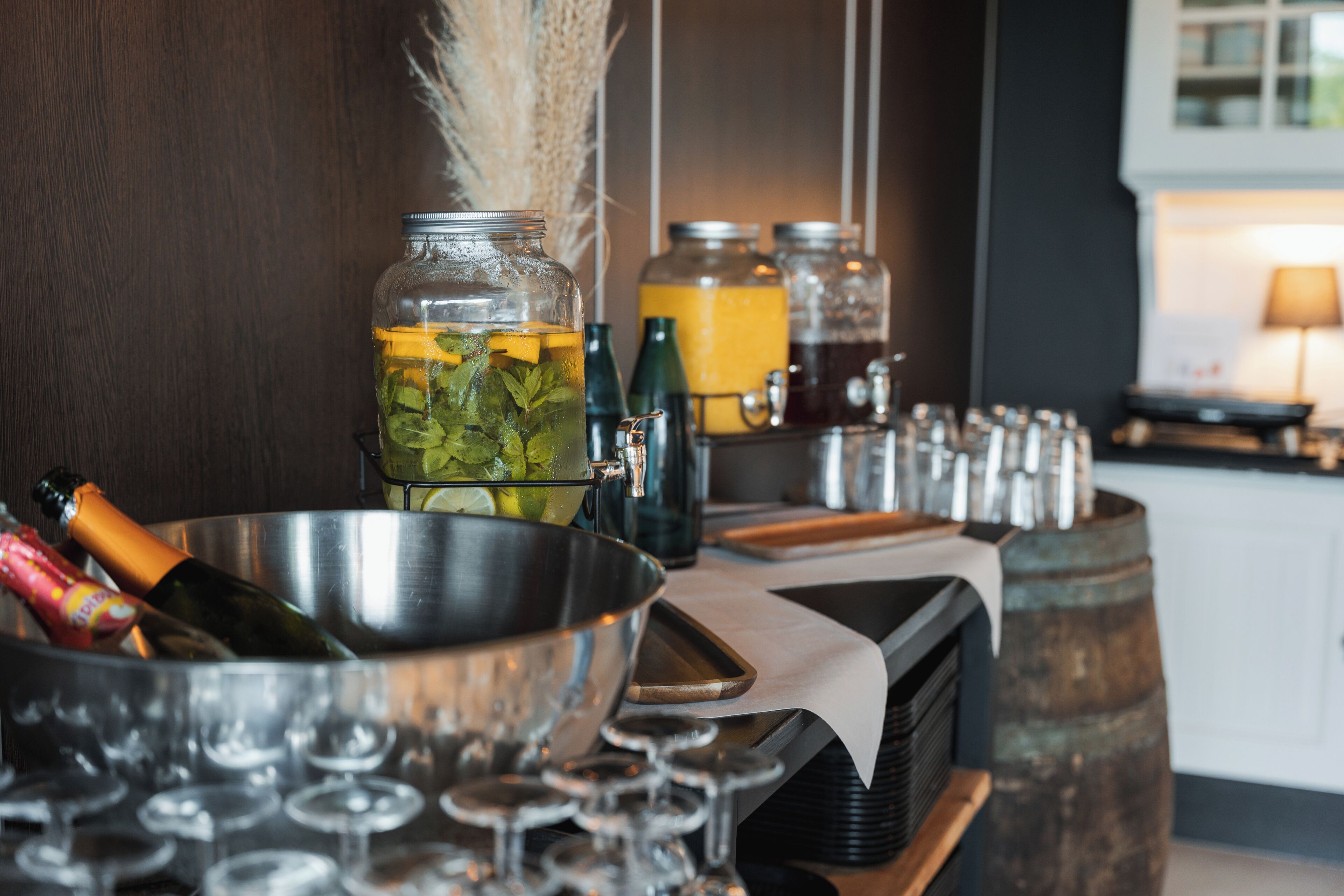 A bar area with a metal bowl, glasses, and bottles, with a wooden backdrop and a kitchen in the background.