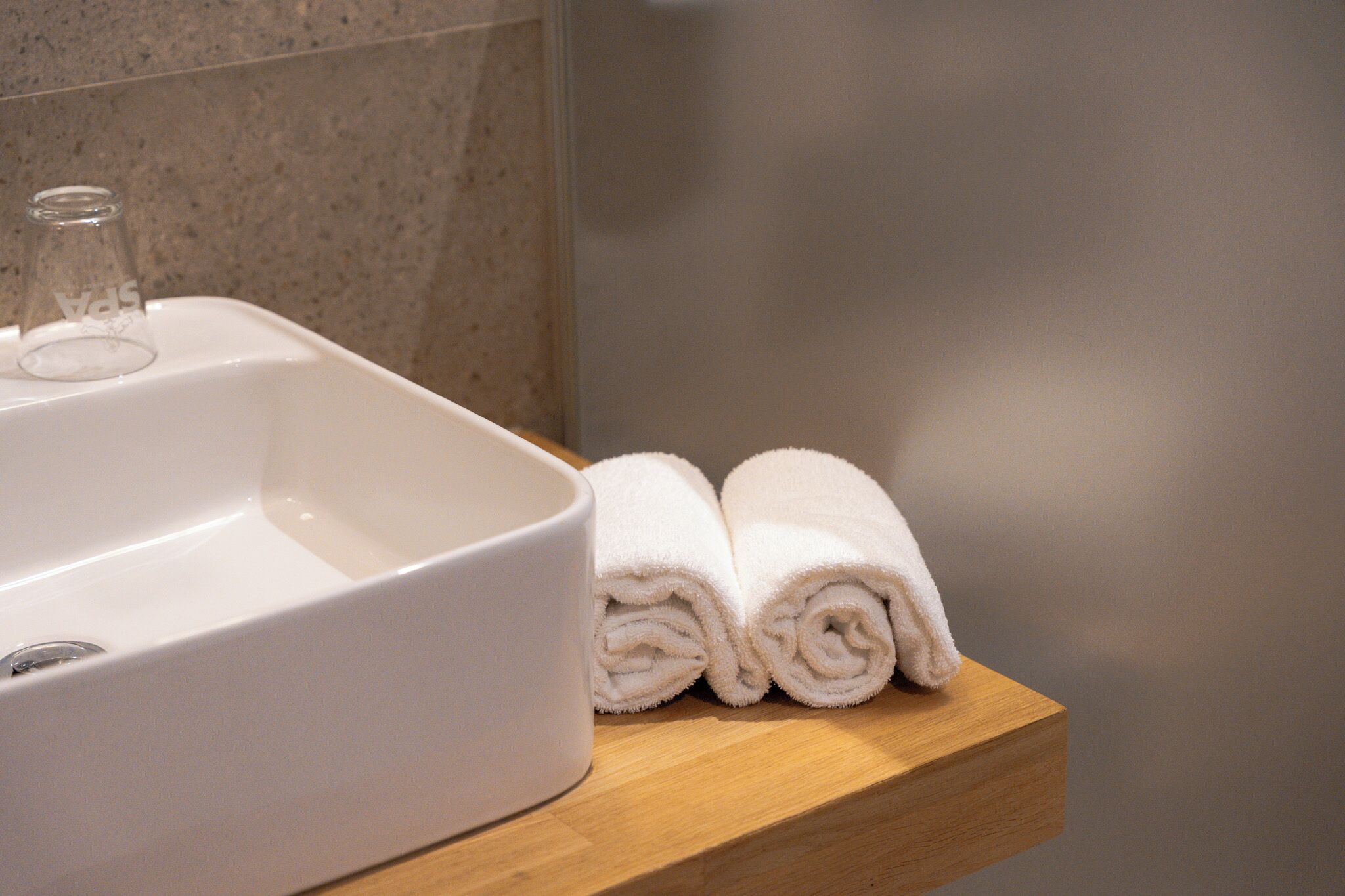 Un baño con un lavamanos blanco, una botella de vidrio y dos toallas enrolladas sobre una repisa de madera.