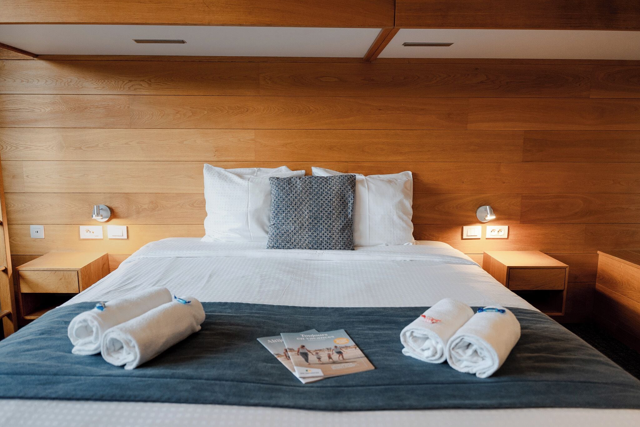 Une chambre d’hôtel avec une tête de lit en bois, deux tables de chevet avec des lampes, un lit avec du linge de lit blanc et des serviettes roulées posées sur le lit.