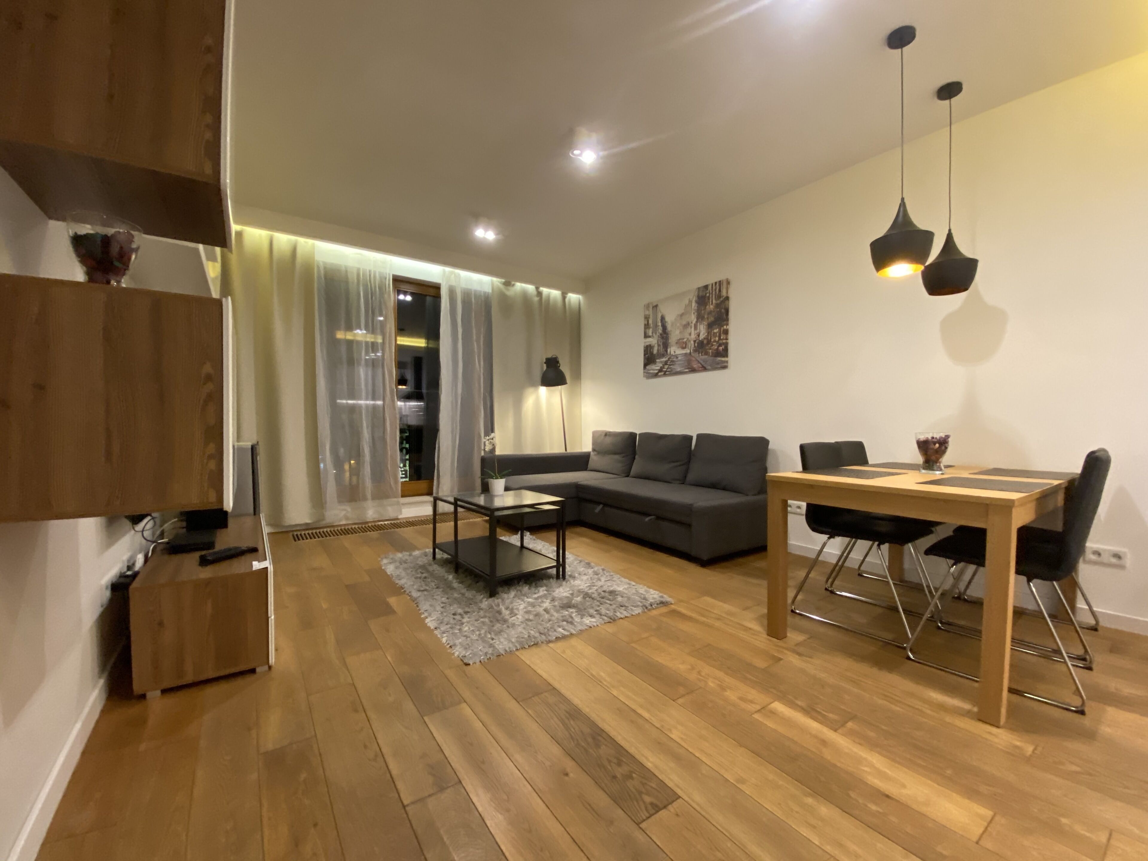 A modern living room with a sofa, dining table, and chairs. There is a wooden cabinet, a small table with a lamp, and a painting on the wall.