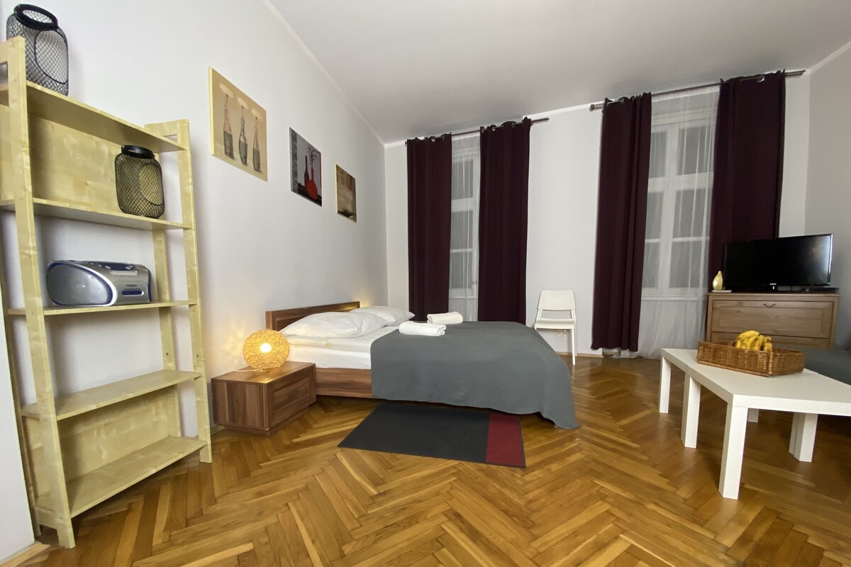 A hotel room with a wooden herringbone floor, a bed with white linens, a bedside table with a lamp, a TV on a wooden cabinet, and a wicker basket on a side table.