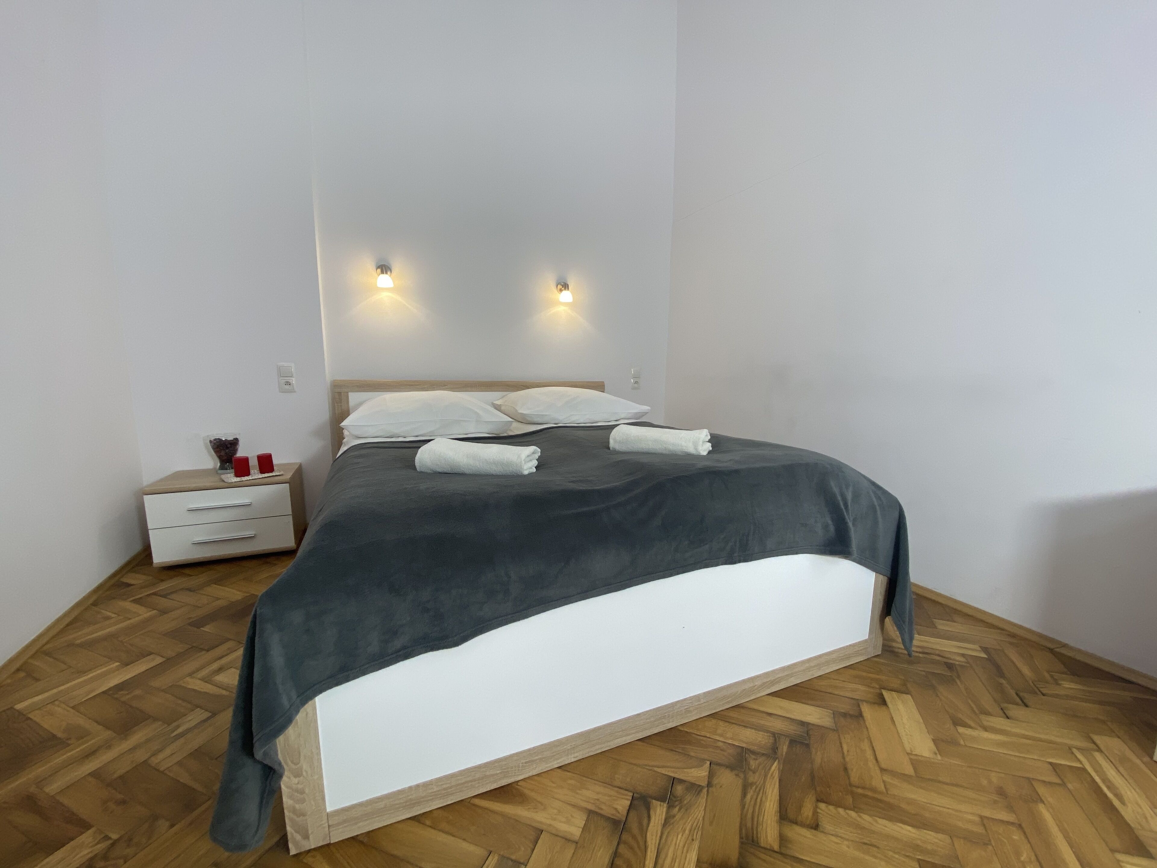 A neatly made bed with a grey bedspread and white pillows, a wooden nightstand with a few items on it, and two wall-mounted lights.