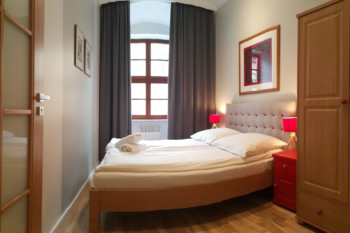 A neatly made bed with pillows and a towel, a bedside table with a lamp, a wardrobe, and a window with curtains.