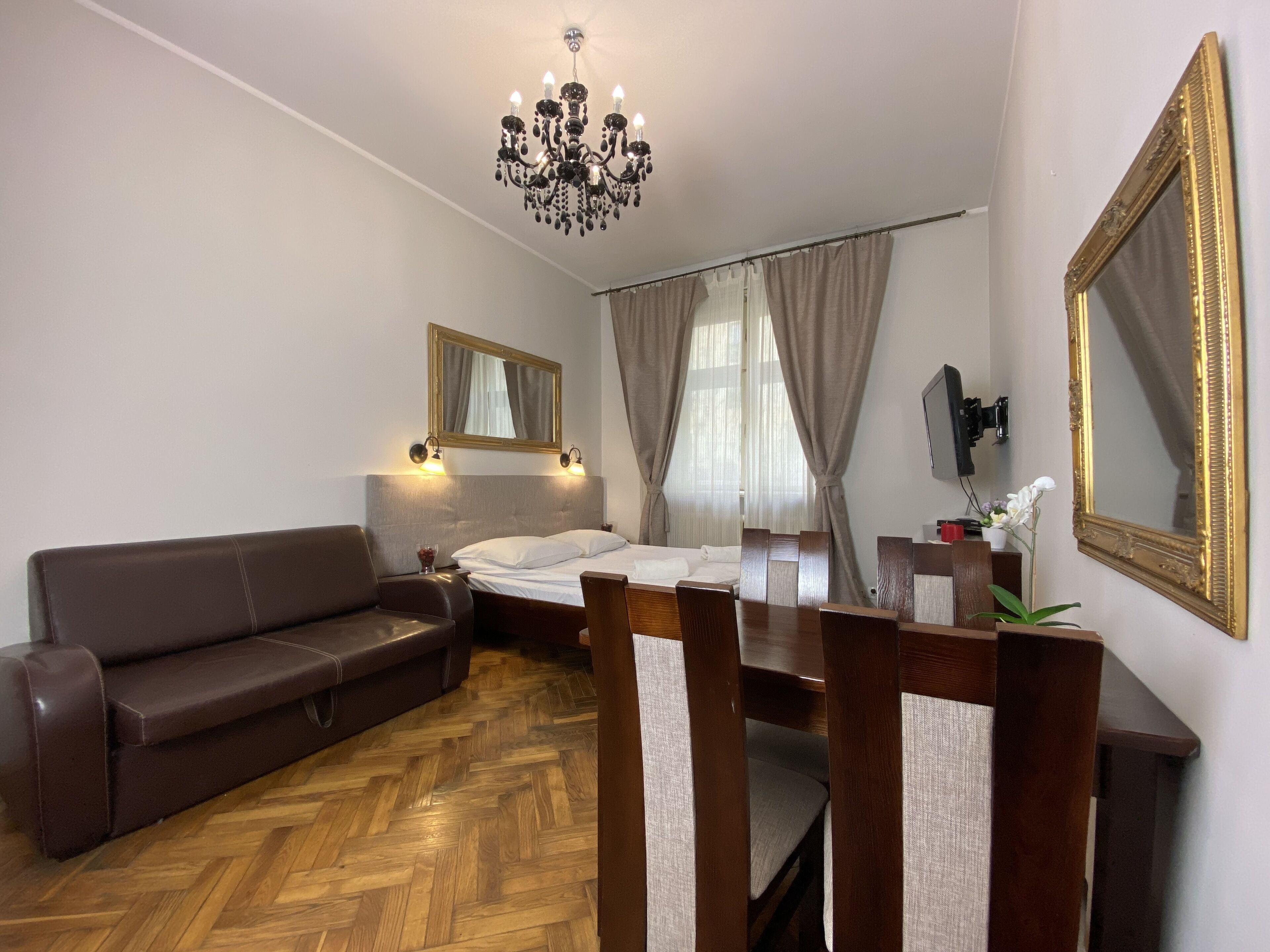 Quarto de hotel com chão de madeira em padrão herringbone, sofá de couro marrom, mesa de jantar de madeira com cadeiras, cama com roupa de cama branca, lustre e um espelho grande com moldura dourada.