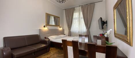 Quarto de hotel com chão de madeira em padrão herringbone, sofá de couro marrom, mesa de jantar de madeira com cadeiras, cama com roupa de cama branca, lustre e um espelho grande com moldura dourada.