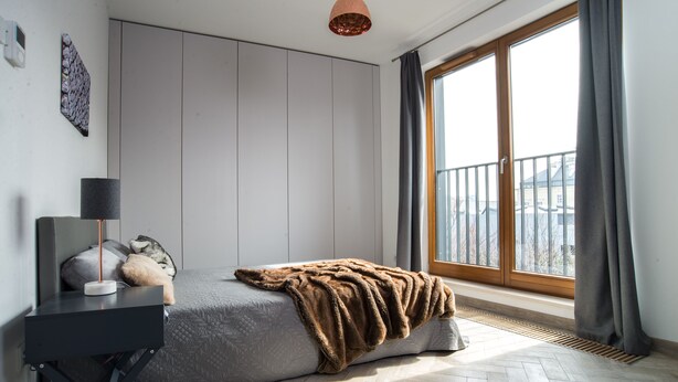 A modern bedroom with a bed, a bedside table, a lamp, and a view through a large window. - P&J Apartments (Kraków)