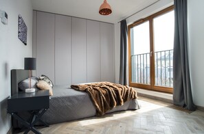A modern bedroom with a bed, bedside table, lamp, and a view of the city through large windows. - P&J Apartments (Kraków)