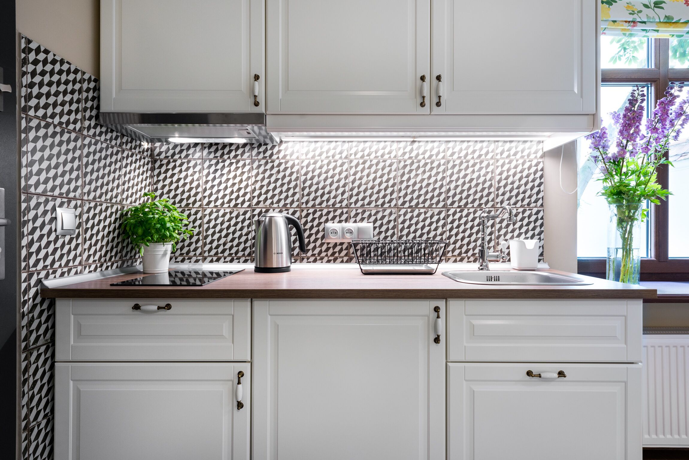 A kitchen with white cabinets, a patterned backsplash, a kettle, a coffee maker, and a potted plant.