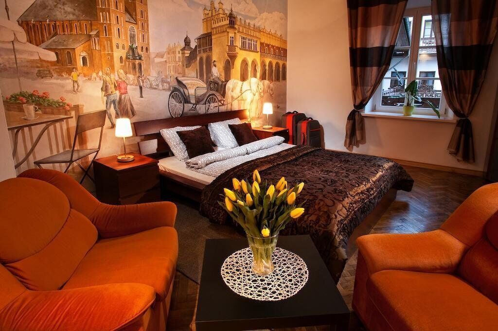 Ein Hotelzimmer mit einem Bett, zwei orangefarbenen Stühlen, einem Nachttisch mit Lampe sowie einer Vase mit Blumen und einem Wandgemälde eines historischen Gebäudes.