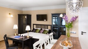 A hotel room with a bed, a desk with two lamps, a dining table with chairs, a chandelier, and a vase with flowers. - P&J Apartments (Kraków)