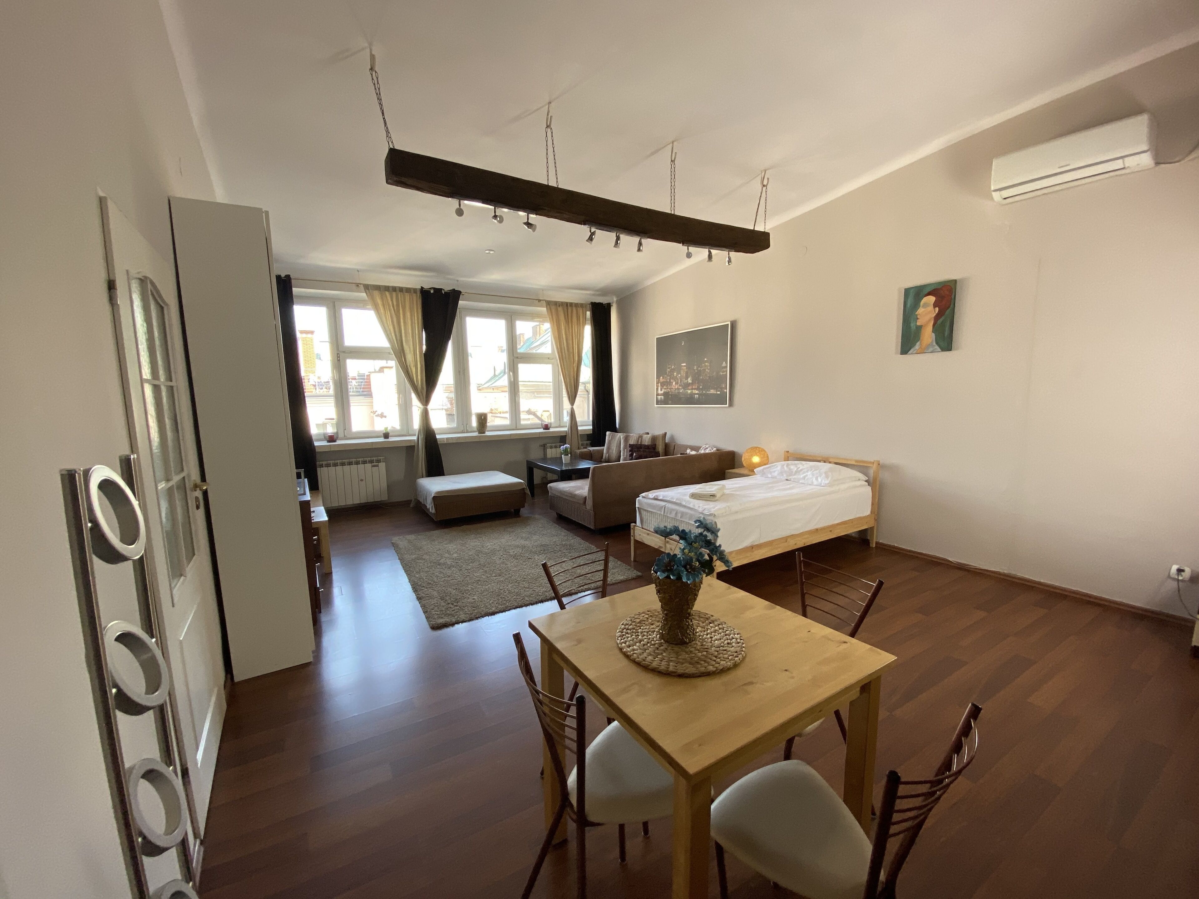 A compact living space with a bed, a sofa, a dining table with chairs, a small kitchen area, and a large window with curtains.