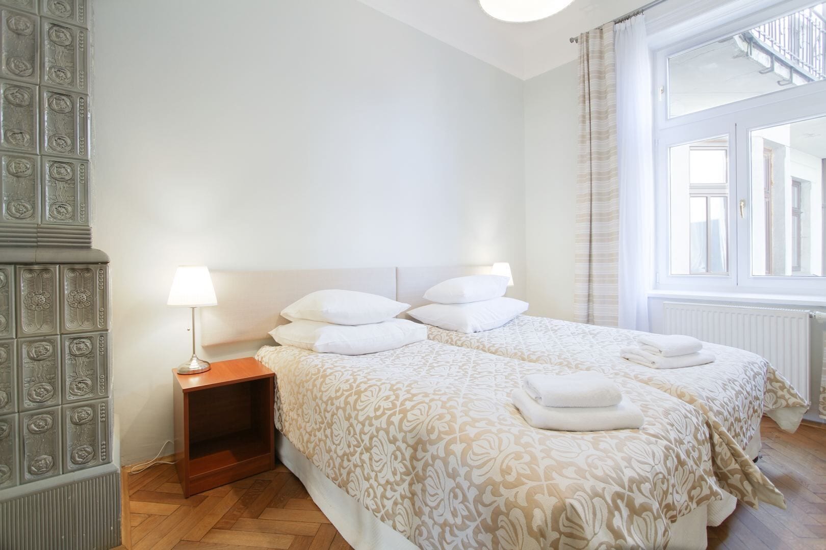 A hotel room with two beds, a nightstand, a lamp, and a view of the outside through a window.