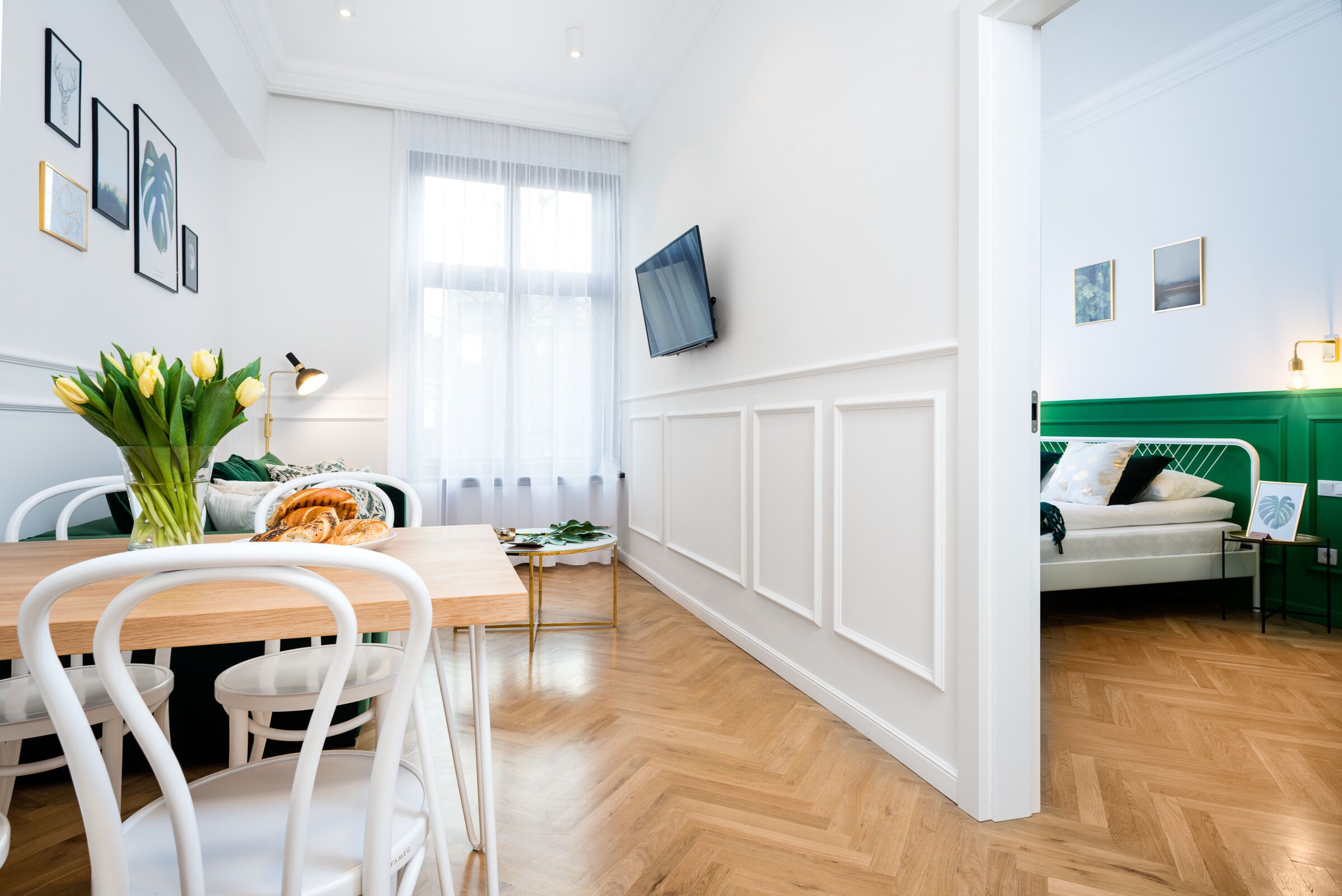 A modern living room with a wooden dining table, white chairs, a sofa, and a TV mounted on the wall.