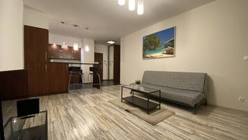 Una sala de estar moderna con un sofá gris, una mesa de centro y un mueble para TV con control remoto. Se aprecia una zona de cocina con armarios de madera y una mesa de comedor con dos sillas.