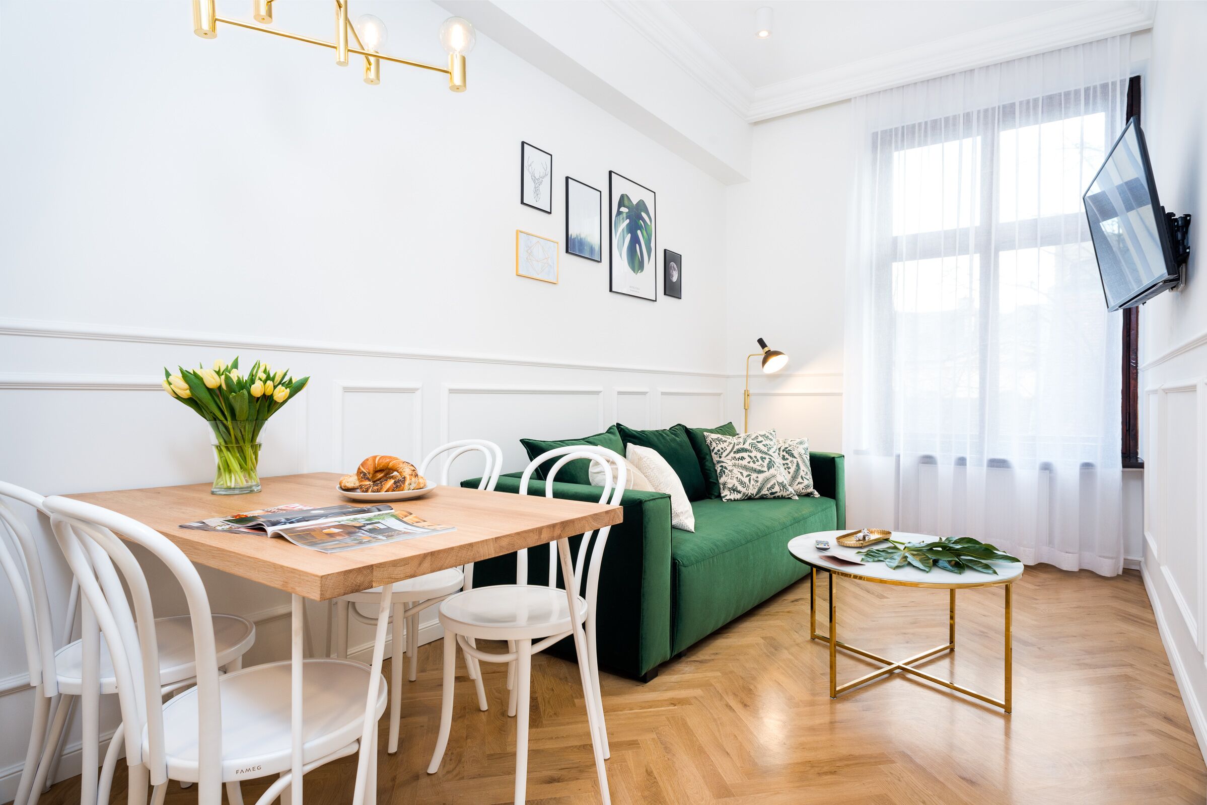 A modern living room with a wooden dining table, white chairs, a green sofa, and a small round table with a lamp.