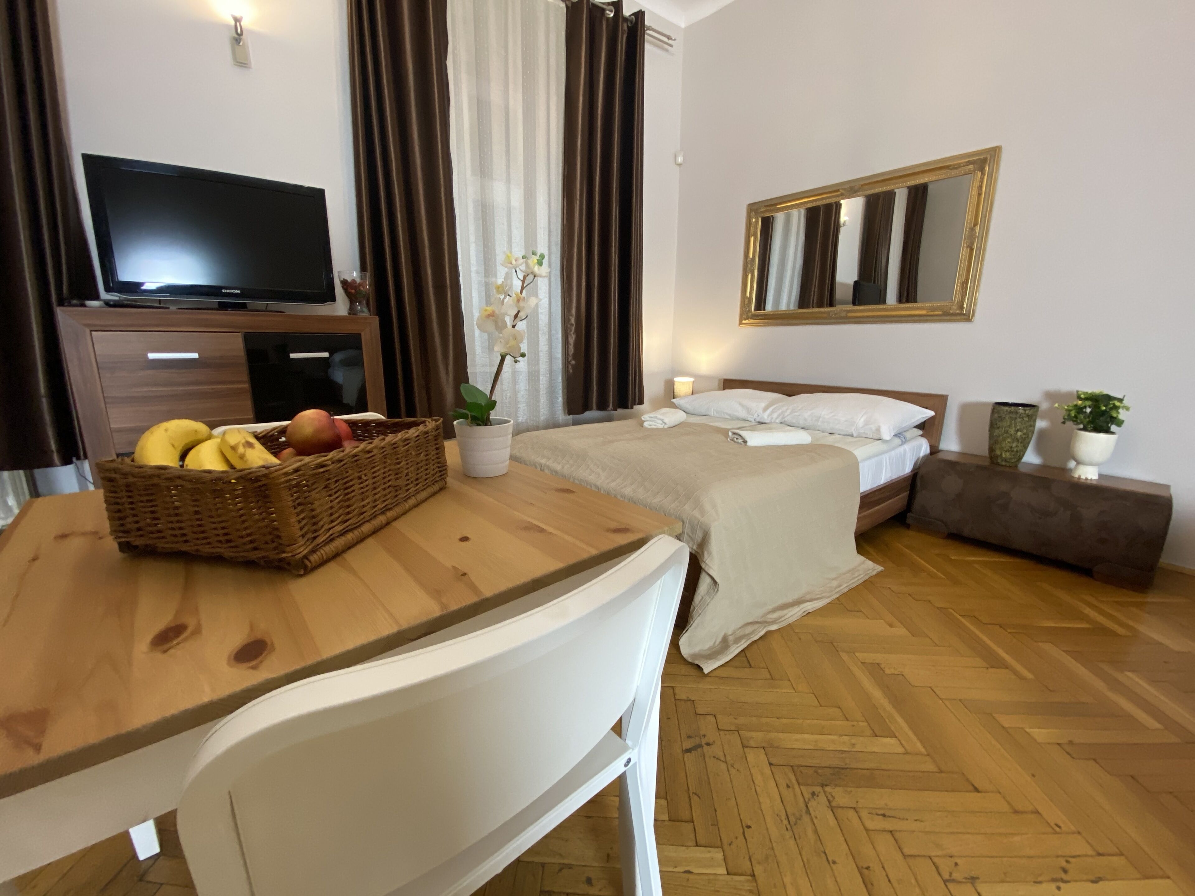 A hotel room with a wooden desk, a television, a bed with white bedding, a mirror, and a potted plant.