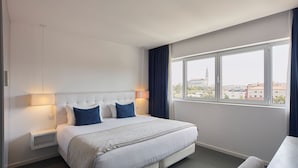A hotel room with a large bed, a bedside table, a lamp, a window with a city view, and a chair.