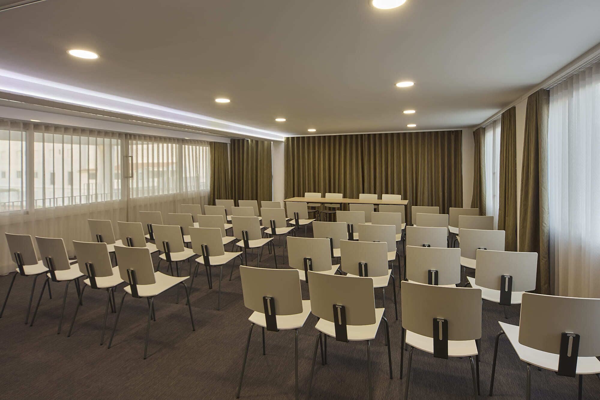 Un salón de conferencias con filas de sillas, una mesa larga y ventanas grandes con cortinas.