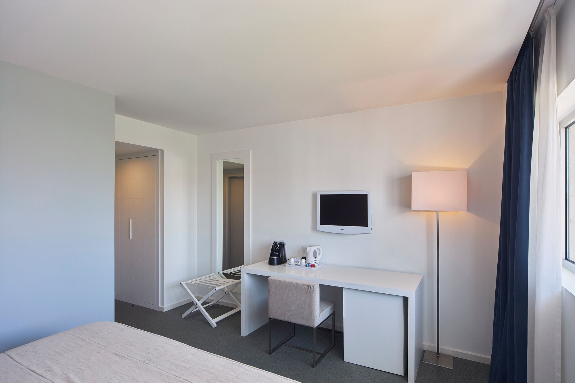 A hotel room with a bed, a desk with a TV, a lamp, and a clothes drying rack.