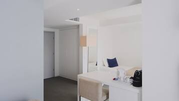 A hotel room with a white desk, chair, and a bed with a blue pillow. There is a coffee maker and a bag on the desk.
