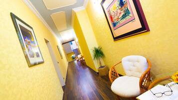 A hallway with wooden flooring, yellow walls, and framed artwork. There is a wicker chair and a table with glasses and a notepad.