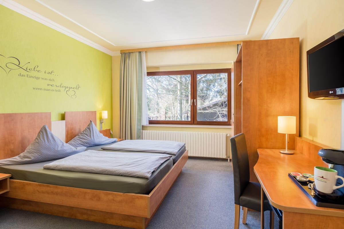 A hotel room with a bed, a desk with a coffee machine, a TV, and a window with a view of trees.