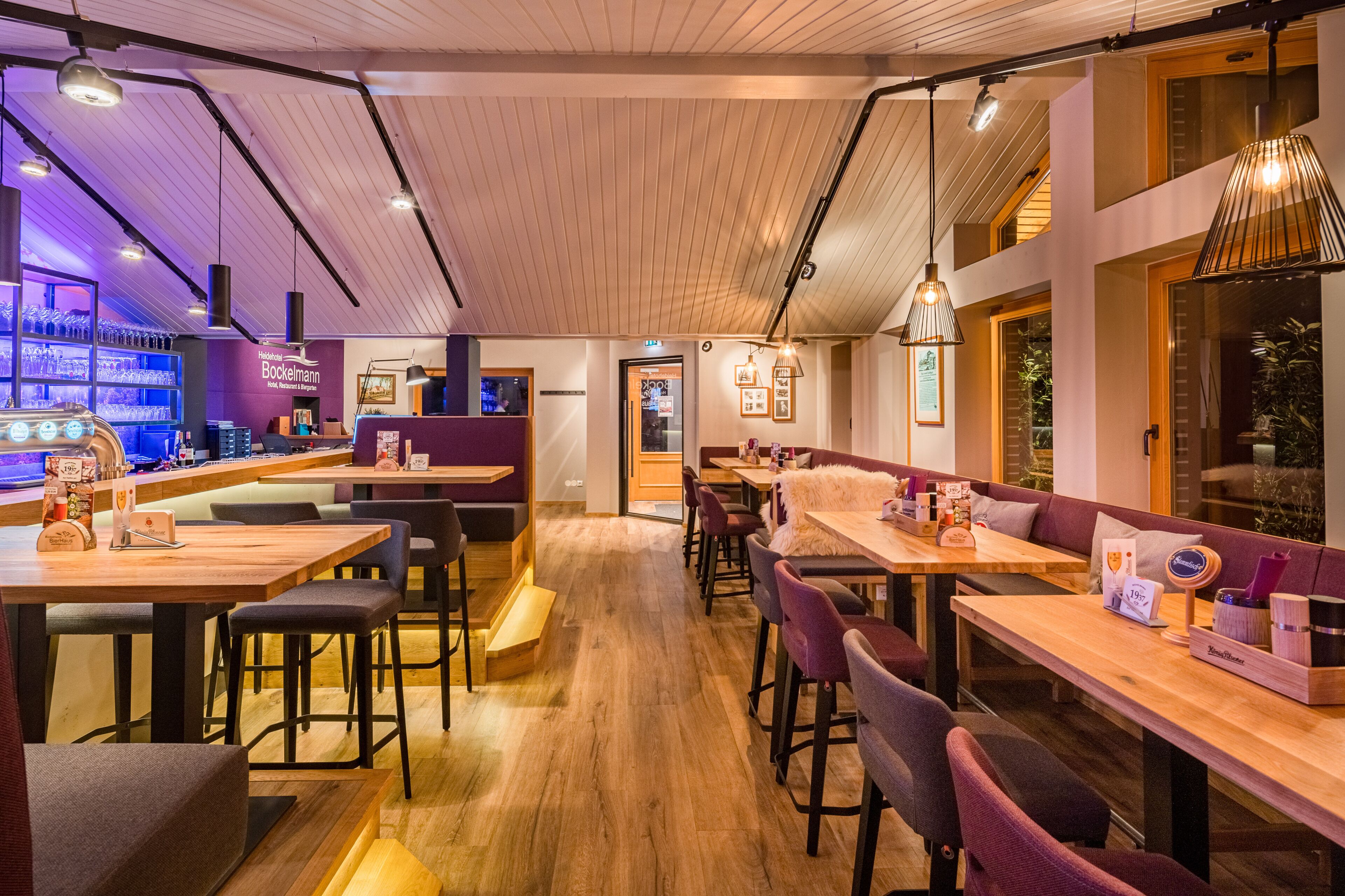 A restaurant with wooden tables and chairs, a bar area, and pendant lighting.