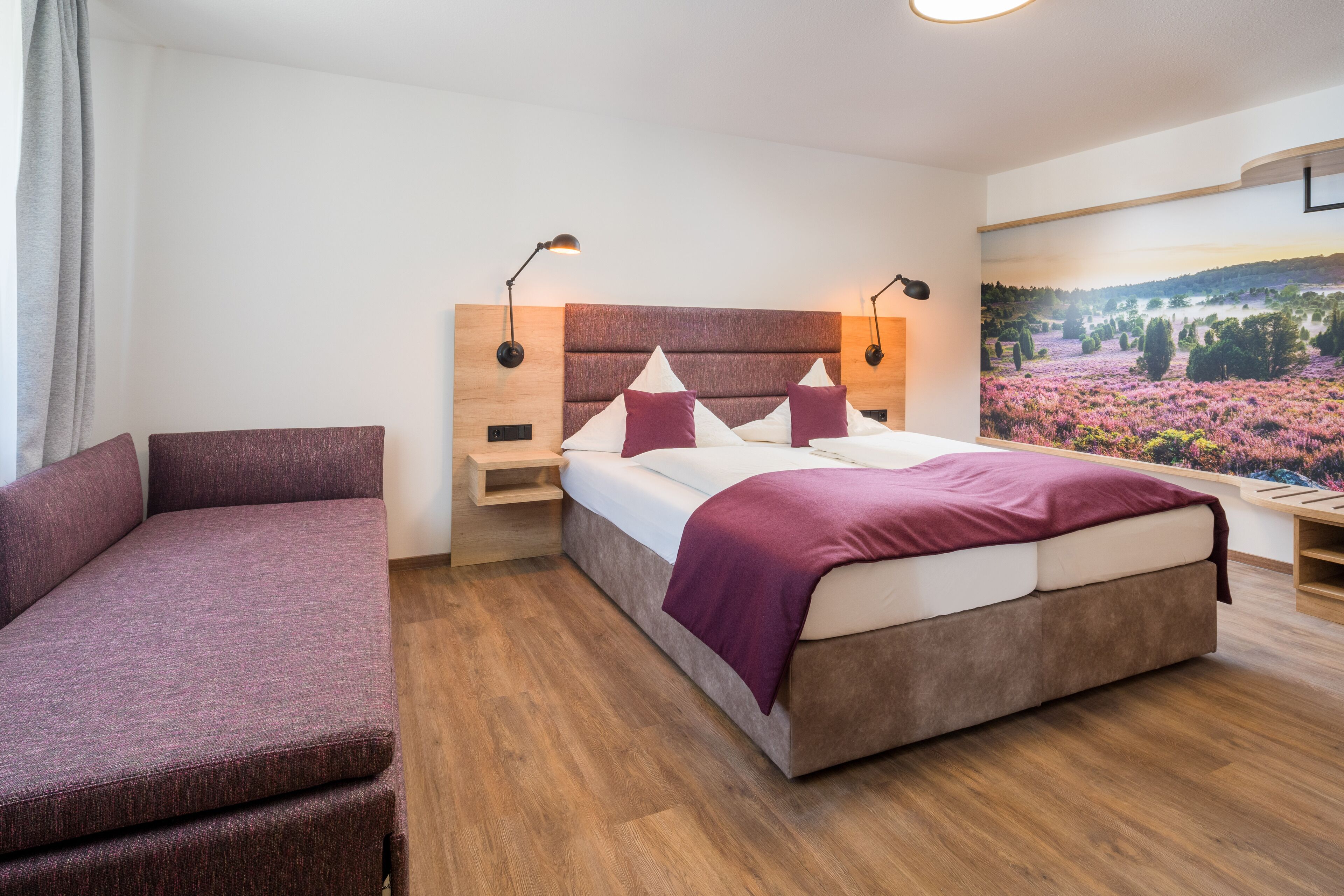 A modern hotel room with a bed, a sofa, and a large landscape painting on the wall.