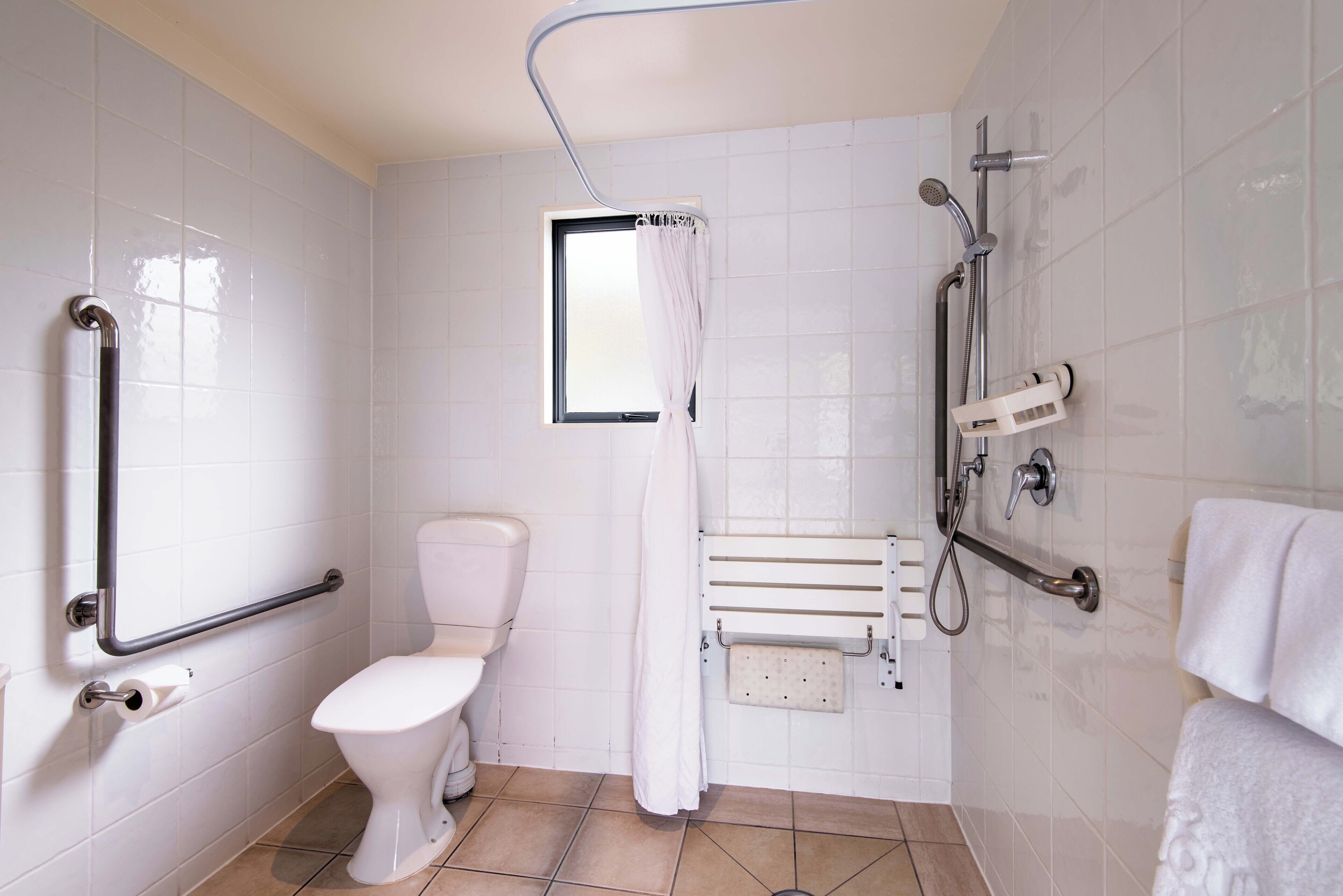A bathroom with a toilet, a shower area with a handheld showerhead, a grab bar, and a window with a curtain.