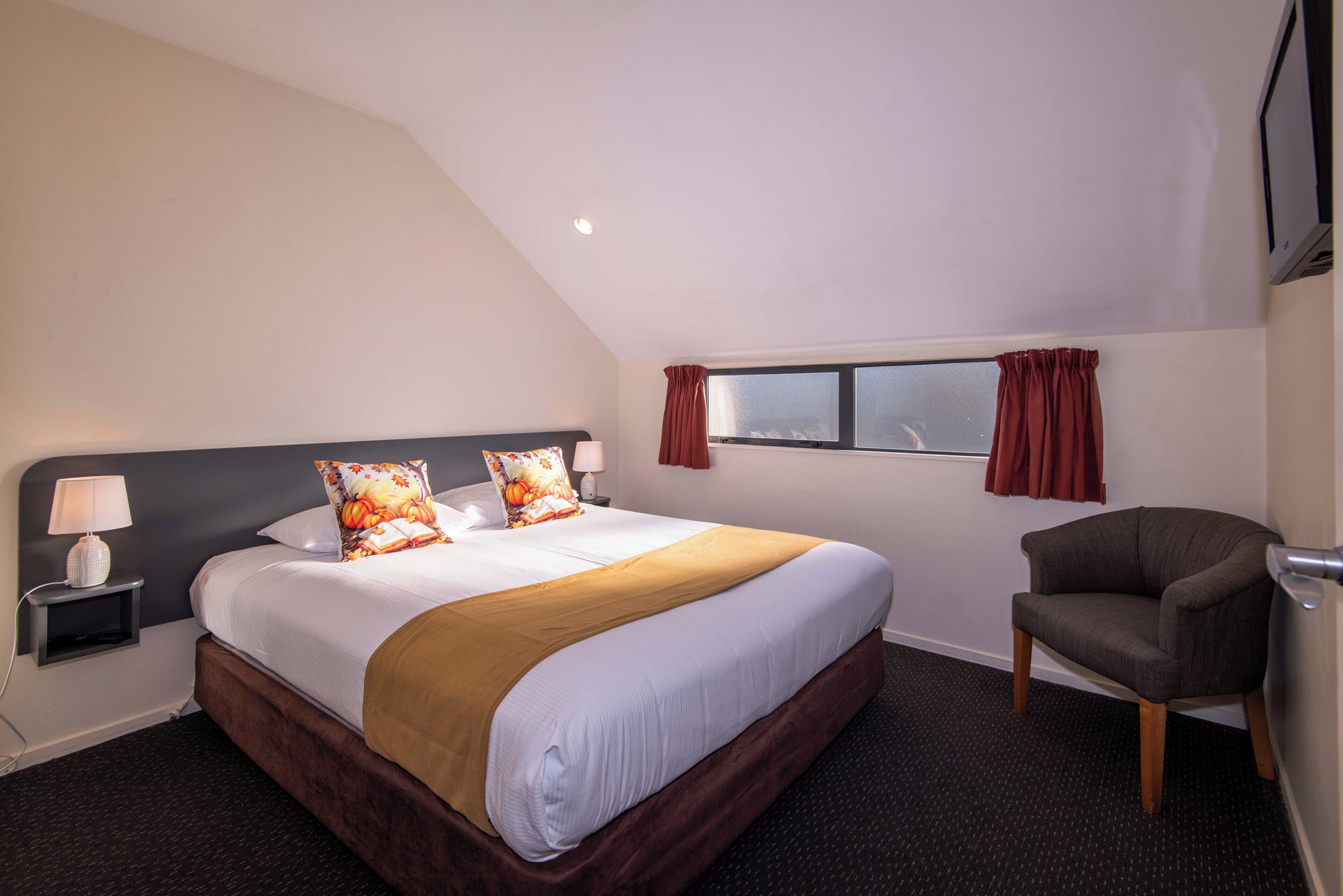 Une chambre d’hôtel avec un grand lit, une chaise, une table de chevet avec une lampe, une fenêtre avec des rideaux et un téléviseur fixé au mur.