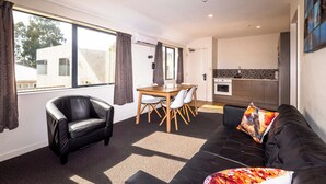 A modern living room with a black leather sofa, a dining table with chairs, and a kitchen area visible through an open door. - 298 Westside Motor Lodge (Christchurch)