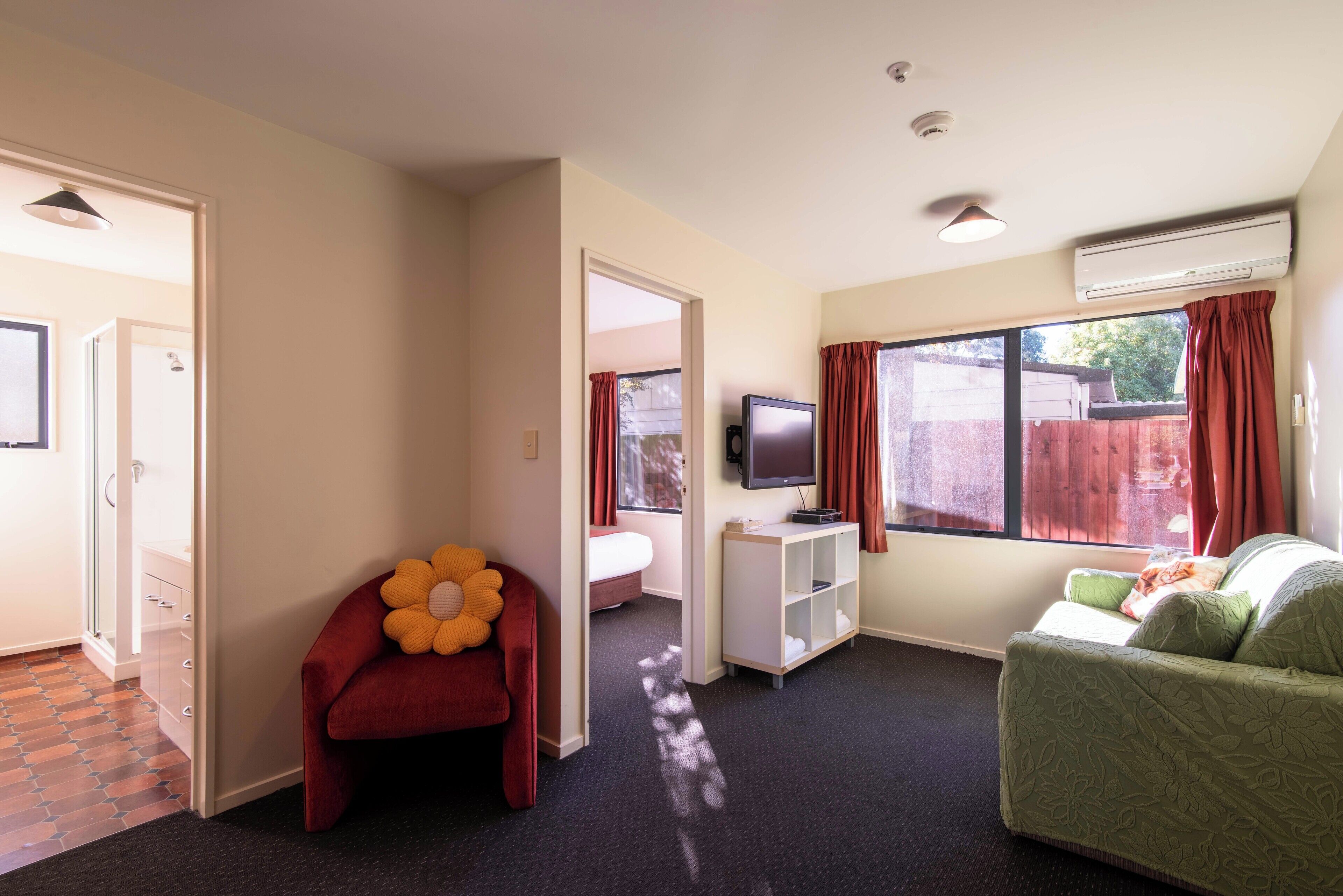 A hotel room with a bed, a TV, a red chair, a green sofa, and a window with curtains.