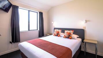 Une chambre d’hôtel dotée d’un grand lit, d’un téléviseur à écran plat fixé au mur, d’une fenêtre avec des rideaux et d’une table de chevet avec une lampe.