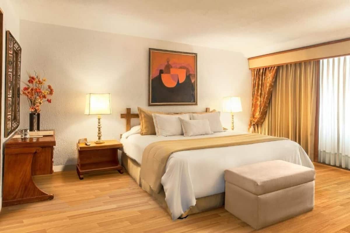 A hotel room with a bed, two bedside tables, a nightstand, a lamp, a painting, and a window with curtains.