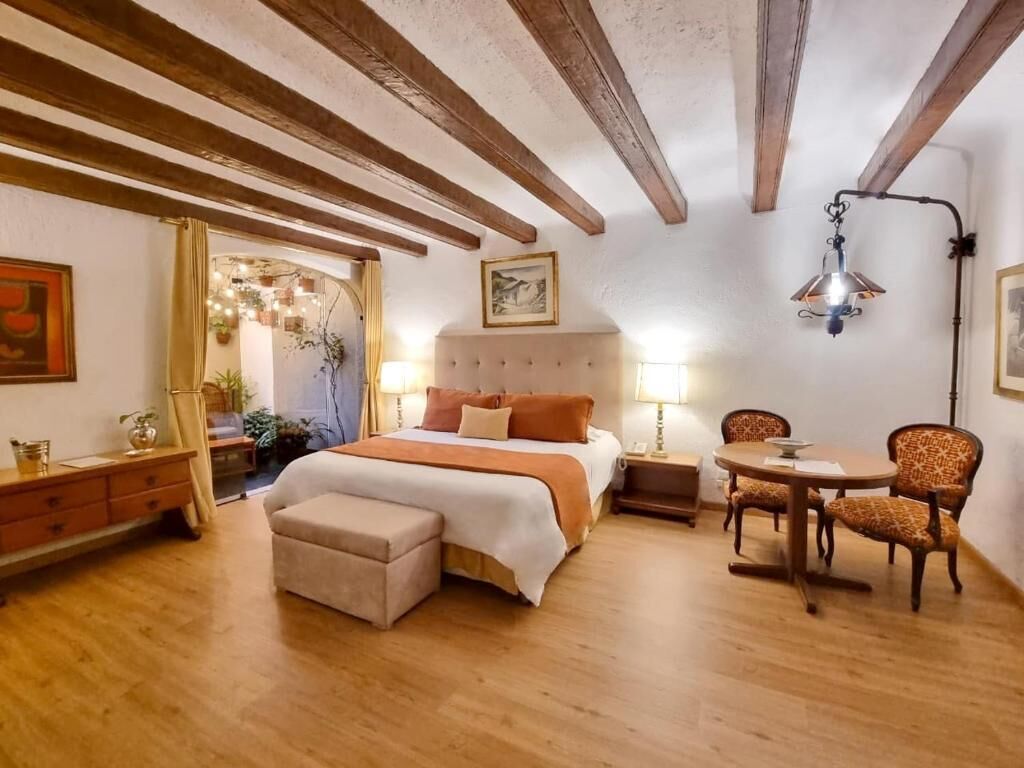 A bedroom with a bed, bedside table, lamp, and a dining area with chairs.