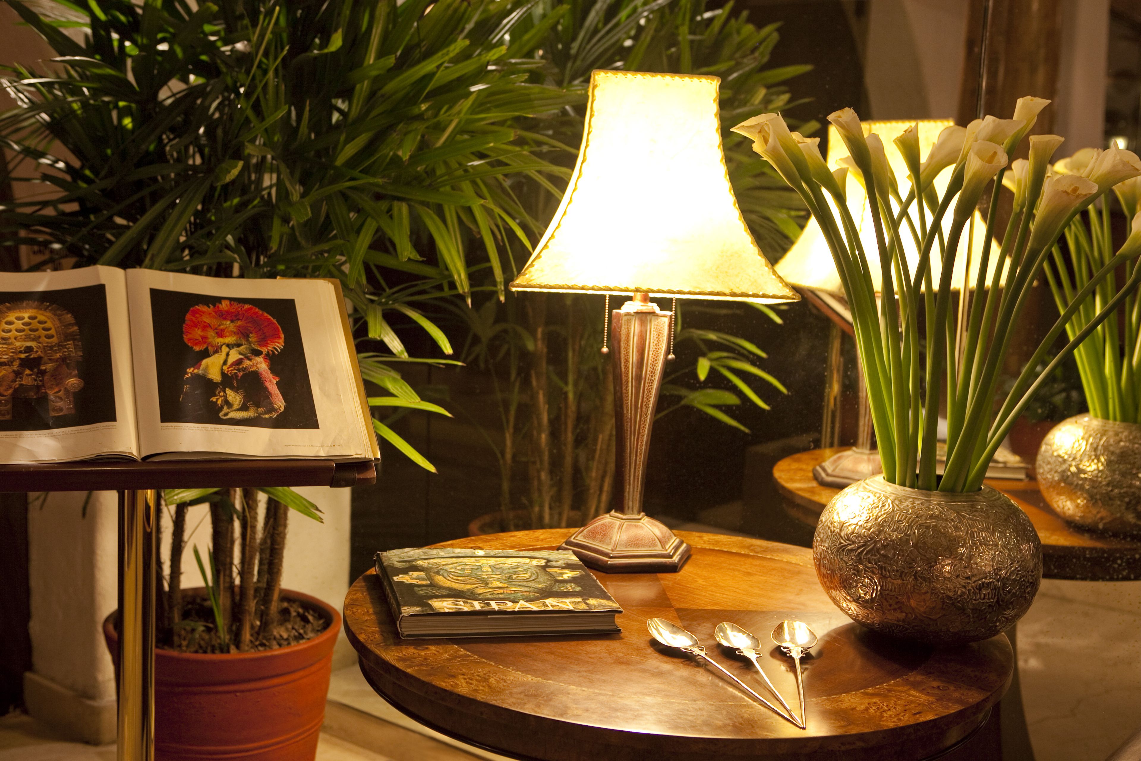A round wooden table with a book, a lamp, a vase with flowers, and a potted plant.
