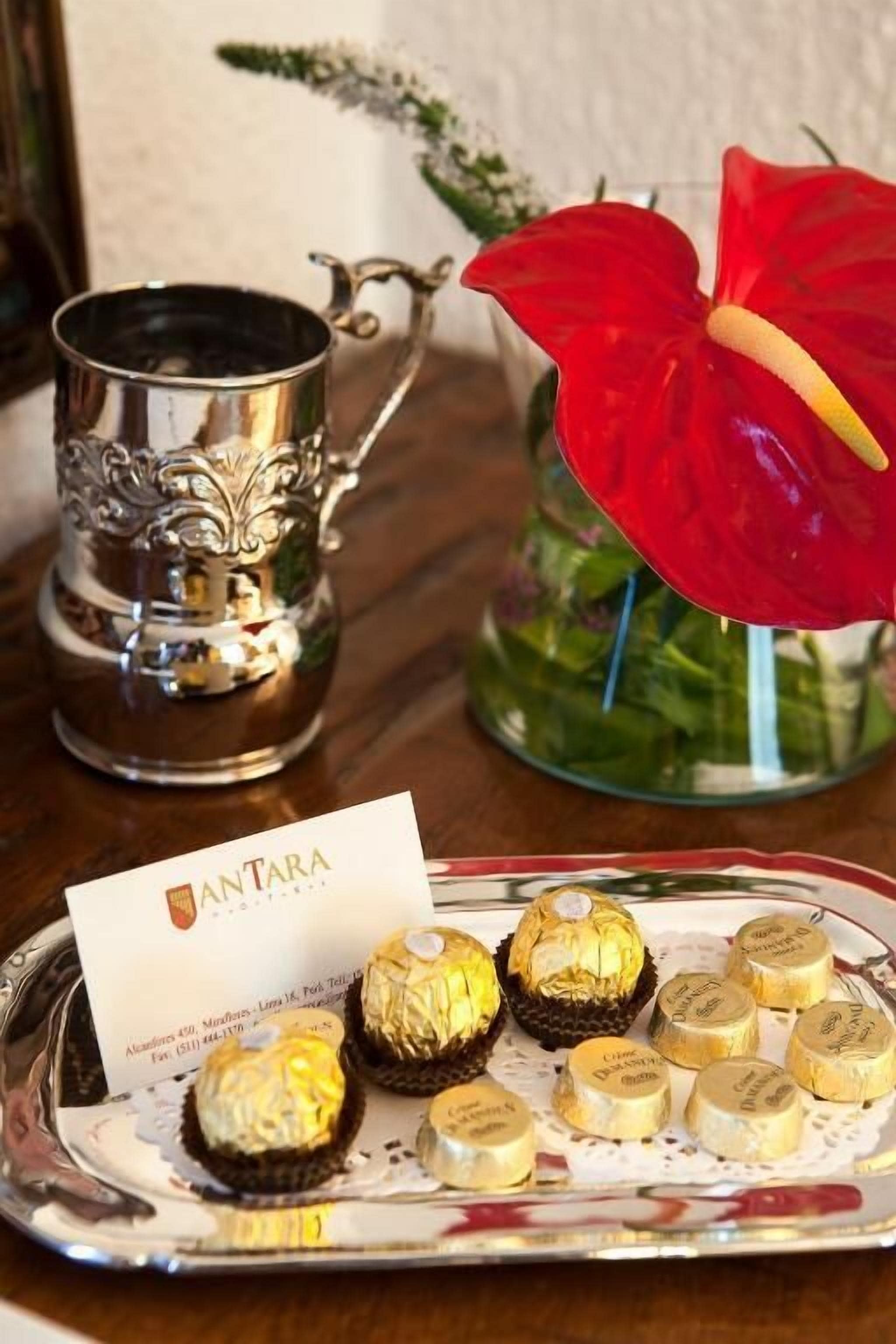 Un plateau contenant un assortiment de chocolats et une tasse en argent, une fleur rouge et une carte de visite, le tout posé sur une table en bois.