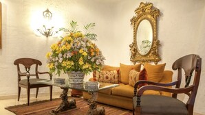 Un salon comprenant un canapé, deux fauteuils, une table basse en verre agrémentée d’un arrangement floral, un tapis à motifs et un miroir décoratif accroché au mur.