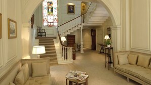 A spacious living room with a staircase leading to a second floor, a comfortable sofa, a small table with magazines, and a decorative stained glass window. - Eshott Hall (Morpeth)