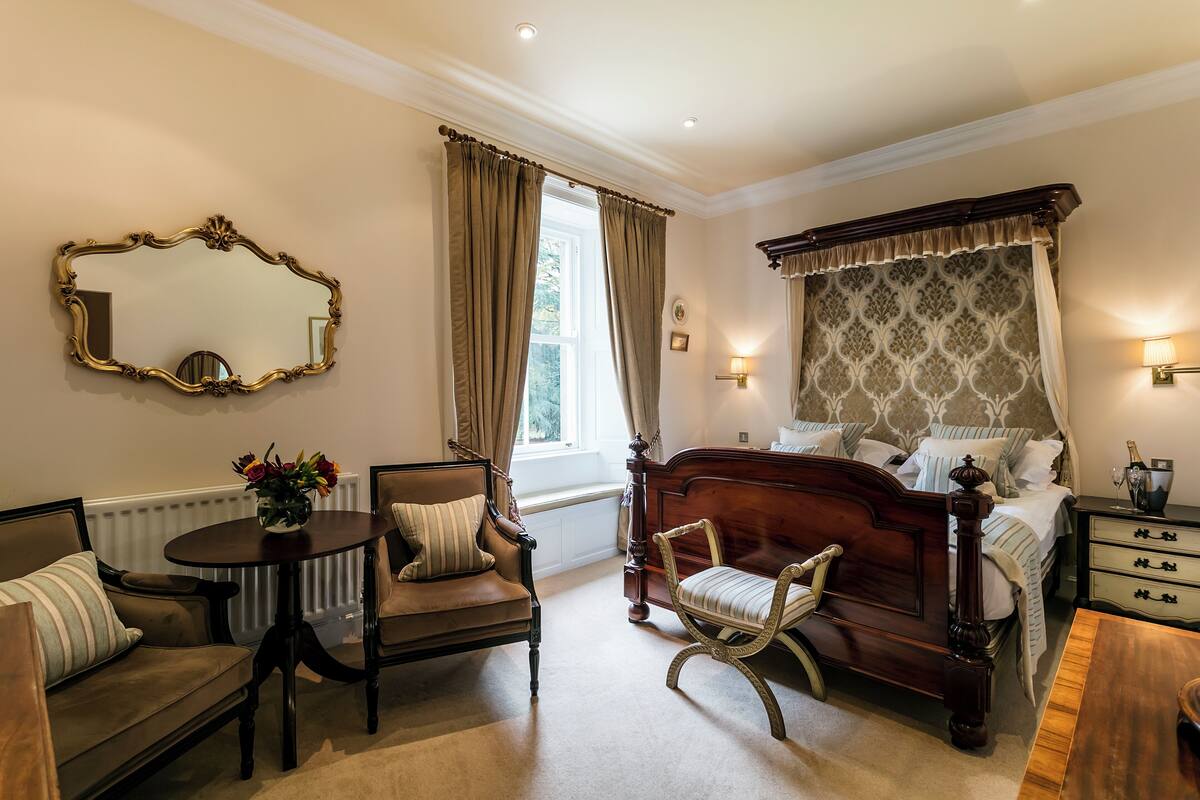 Quarto com cama grande, área de estar com poltrona e mesa, espelho e janela com cortinas.